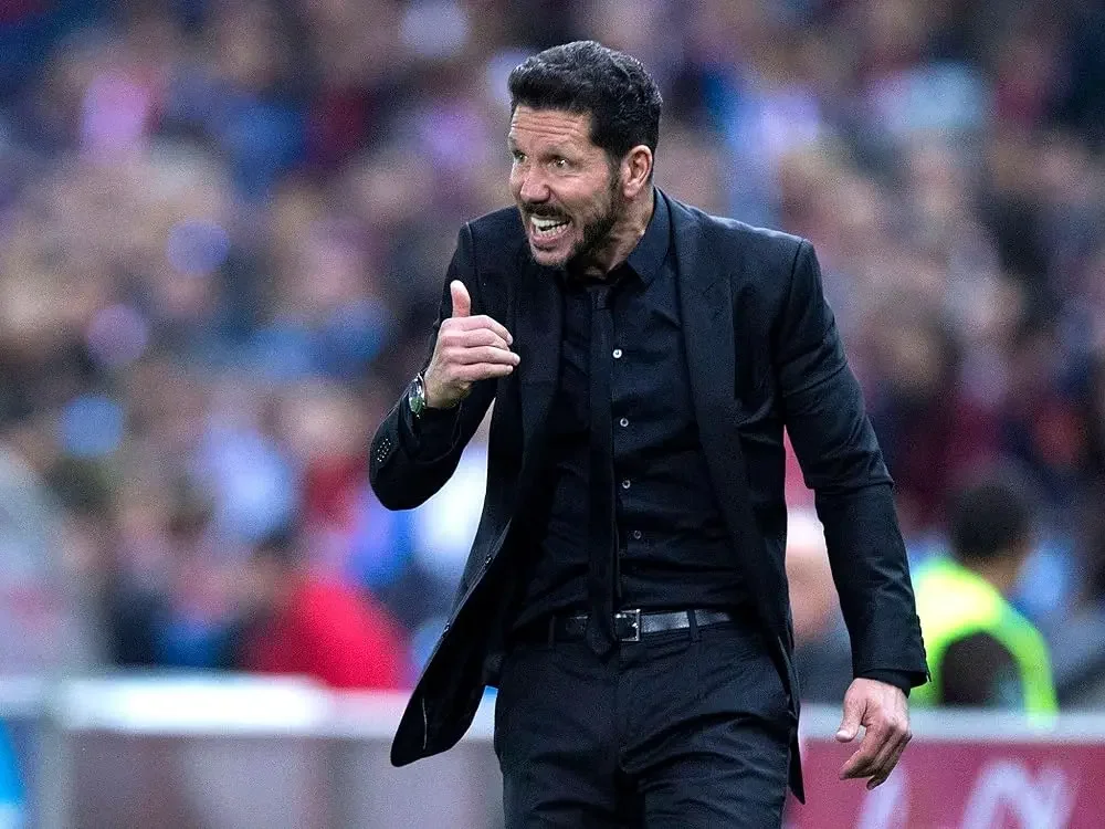 Diego Simeone wearing a black suit and shirt, gesturing emotionally during a football match.